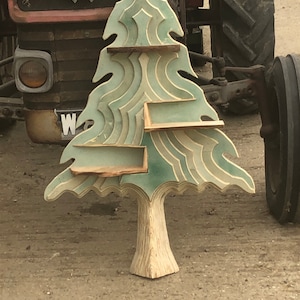 May include: A wooden tree-shaped shelf with three shelves. The shelves are painted in shades of green and white. The tree is standing on a brown surface.