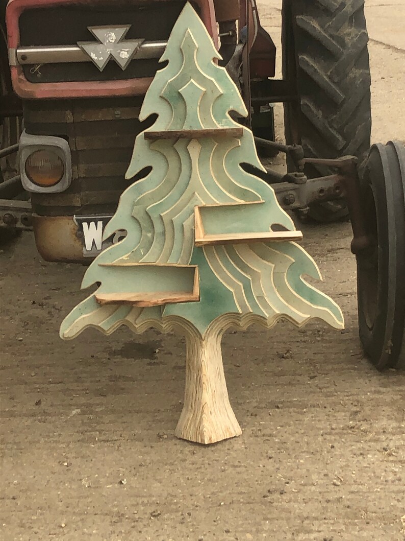 May include: A wooden tree-shaped shelf with three shelves. The shelves are painted in shades of green and white. The tree is standing on a brown surface.
