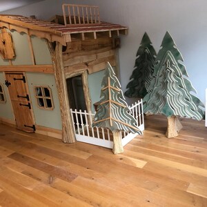 May include: A wooden playhouse with a green exterior, a brown door, and two windows. The playhouse has a balcony and a white picket fence. There are three green wooden trees and a white picket fence in the background.