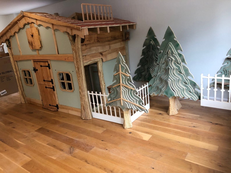 May include: A wooden playhouse with a green exterior, a brown door, and two windows. The playhouse has a balcony and a white picket fence. There are three green wooden trees and a white picket fence in the background.