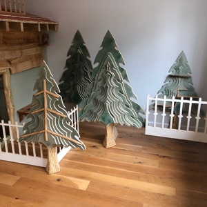 May include: A playroom featuring a wooden playhouse and three large green wooden trees. The trees are crafted from layered wood, creating a 3D effect. A white picket fence stands in front of the playhouse, with a white gate in the corner of the room.