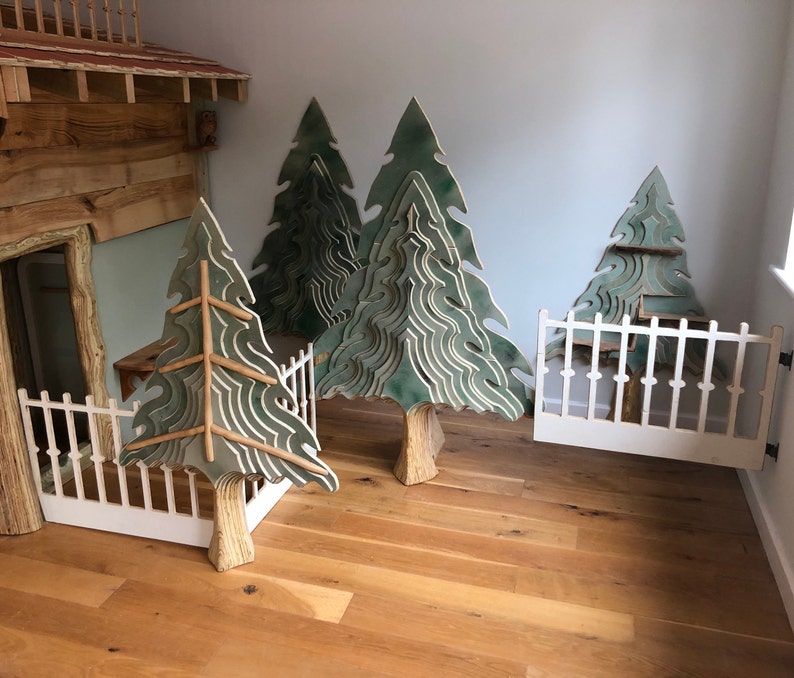 May include: A playroom featuring a wooden playhouse and three large green wooden trees. The trees are crafted from layered wood, creating a 3D effect. A white picket fence stands in front of the playhouse, with a white gate in the corner of the room.