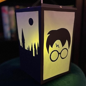 May include: A yellow and blue paper lantern with a silhouette of Hogwarts castle on one side and a silhouette of Harry Potter on the other side. The lantern is lit from inside, casting a warm glow.