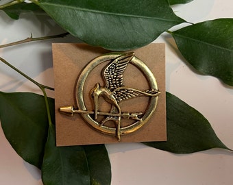 Pode incluir: Um alfinete dourado com um pássaro com as asas abertas, pousado em uma flecha dentro de uma moldura circular. O alfinete está em um pequeno cartão quadrado marrom. Folhas verdes no fundo.