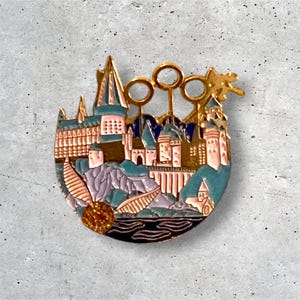 K&ouml;nnte beinhalten: Eine goldfarbene Emaille-Anstecknadel, die Hogwarts Castle darstellt, die legend&auml;re Schule f&uuml;r Hexerei und Zauberei aus der Harry-Potter-Reihe. Die Anstecknadel zeigt eine detaillierte Darstellung des Schlosses, einschlie&szlig;lich seiner T&uuml;rme, Fenster und des Gel&auml;ndes. Die Anstecknadel enth&auml;lt auch eine Darstellung des Goldenen Schnitzers, eines magischen Objekts, das im Sport Quidditch verwendet wird.