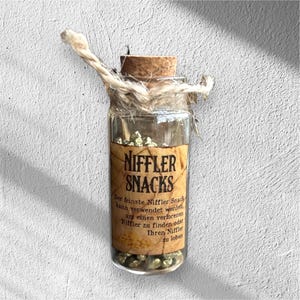 Könnte beinhalten: Kleine Glasflasche mit Korken, gefüllt mit goldfarbenen Nuggets. Die Flasche hat ein Etikett mit der Aufschrift "NIFFLER SNACKS" in einer Vintage-Schriftart, mit zusätzlichem Text darunter. Die Flasche ist mit Bindfaden gebunden.