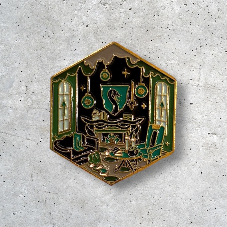 K&ouml;nnte beinhalten: Eine gr&uuml;ne und goldene Emaille-Anstecknadel, die einen gem&uuml;tlichen Raum mit einem Kamin, einem Schlangenwappen und einem gr&uuml;nen Sessel darstellt. Die Anstecknadel hat die Form eines Sechsecks.