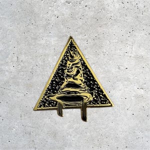 K&ouml;nnte beinhalten: Goldfarbener Metallstift in Form eines Dreiecks mit einem schwarzen und goldenen Zaubererhut im Inneren. Der Hut hat eine spitze Spitze und einen breiten Rand. Der Stift ist ein beliebtes Harry-Potter-Sammlerst&uuml;ck.