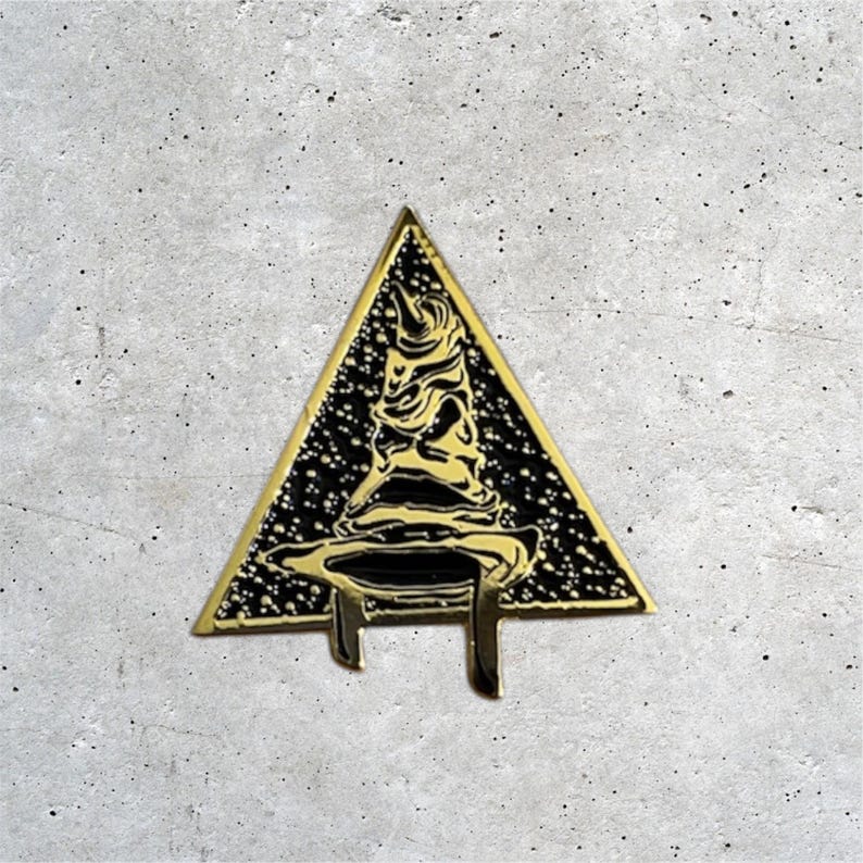 K&ouml;nnte beinhalten: Goldfarbener Metallstift in Form eines Dreiecks mit einem schwarzen und goldenen Zaubererhut im Inneren. Der Hut hat eine spitze Spitze und einen breiten Rand. Der Stift ist ein beliebtes Harry-Potter-Sammlerst&uuml;ck.