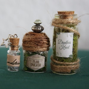 Può includere: Tre piccoli flaconi di vetro con tappi di sughero e spago avvolto attorno al collo. I flaconi contengono erbe essiccate e hanno etichette che recitano "Floh Pulver", "Dianthus Kraut" e "Wendstein".