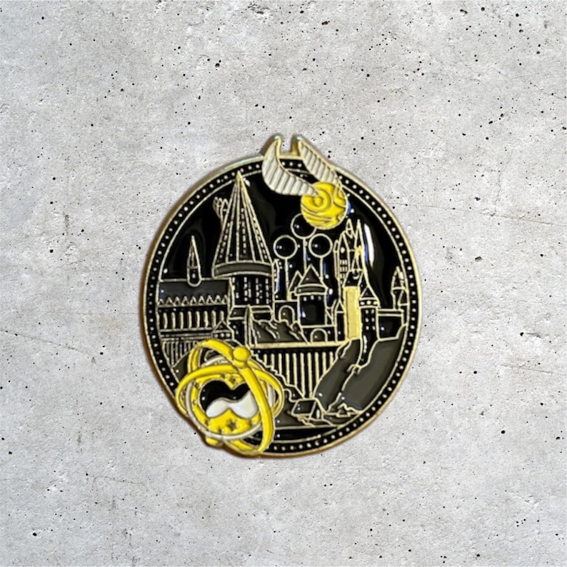 K&ouml;nnte beinhalten: Eine schwarze und goldene Emaille-Anstecknadel mit einer detaillierten Illustration von Hogwarts Castle, dem Goldenen Schnatz und einem Zeitumkehrer. Die Anstecknadel ist rund und hat einen schwarzen Hintergrund.