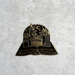 K&ouml;nnte beinhalten: Goldfarbener Emaille-Pin mit einer schwarzen Silhouette von Hogwarts Castle mit einem Halbmond dar&uuml;ber, alles in einem offenen Buch mit einem Symbol der Heiligt&uuml;mer des Todes.
