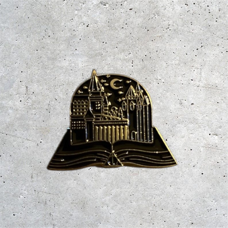 K&ouml;nnte beinhalten: Goldfarbener Emaille-Pin mit einer schwarzen Silhouette von Hogwarts Castle mit einem Halbmond dar&uuml;ber, alles in einem offenen Buch mit einem Symbol der Heiligt&uuml;mer des Todes.