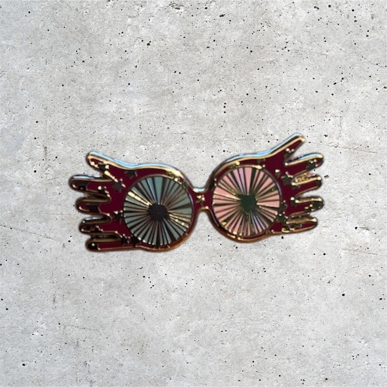 K&ouml;nnte beinhalten: Goldfarbene Emaille-Anstecknadel mit einer Brille mit runden Gl&auml;sern. Die Gl&auml;ser sind mehrfarbig mit einem wirbelnden Muster. Die Brille hat eine einzigartige Form mit einem handf&ouml;rmigen Design an den Seiten.