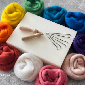 Könnte beinhalten: Ein Filzset mit verschiedenen Farben von Wollvlies, Filznadeln und einer Schaumstoffmatte. Die Farben umfassen Gelb, Orange, Rot, Rosa, Weiß, Blau, Lila und Grün.