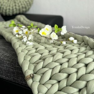 Könnte beinhalten: Eine dicke Strickdecke in Salbeigrün mit gewebter Struktur. Die Decke liegt über einem grauen Sofa mit einem Salbeigrünen Strickkissen im Hintergrund. Die Decke hat ein kleines braunes Leder-Etikett mit dem Wort "TemilaDesign" darauf.