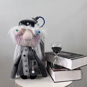 May include: A gray and white plush doll with a large nose and blue eyes. The doll is wearing a gray and white striped jacket and has white hair. The doll is sitting on a table with a book and an hourglass.