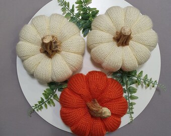 May include: Three knitted pumpkins, two white and one orange, on a white plate with green leaves.