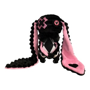 Op de afbeelding: Een zwarte en roze pluche konijn met een zwart-wit geruit patroon op zijn lichaam. Het konijn heeft een roze hart op het ene oor en een zwart kruis op het andere.