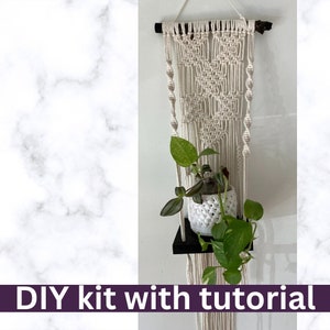 May include: Macrame plant hanger with a black shelf holding a potted plant with green leaves. The macrame is beige and has a geometric design. The text "DIY kit with tutorial" is at the bottom.