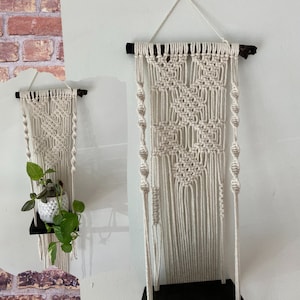 May include: Cream macrame plant hanger with a black shelf. The macrame design features a geometric pattern and hanging strands. The shelf is designed to hold a small potted plant. The hanger is suspended from a natural wooden branch. The shelf is approximately 20 cm wide.