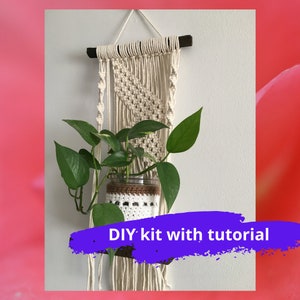 May include: A beige macrame plant hanger with a glass jar containing a green plant. The jar is in a white and brown crocheted sleeve. The macrame hangs from a dark brown wooden rod. Text on the image reads: DIY kit with tutorial.