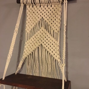 May include: A macrame wall shelf with a wooden shelf and white cotton cords. The shelf is hanging from the wall with two cords on each side. The macrame is in a diamond pattern with fringe at the bottom.
