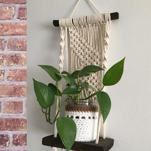 May include: Macrame plant hanger with a dark wooden shelf and a white crocheted pot holding a green leafy plant. The macrame design features a woven pattern and hanging cords. The shelf is approximately 8 inches wide.