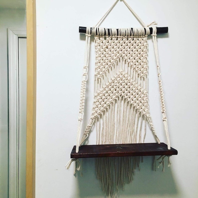 May include: A macrame wall hanging with a wooden shelf. The macrame is made with natural white cotton cord and has a geometric pattern. The shelf is dark brown wood.