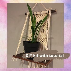 May include: A DIY macrame plant hanger kit with a wooden shelf and a tutorial. The kit includes white macrame cord, a wooden dowel, and a shelf. The kit is shown with a potted plant.