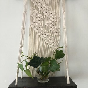 Puede incluir: Un tapiz de macramé de pared con un patrón geométrico, que incluye una estantería de madera con una pequeña planta en maceta.