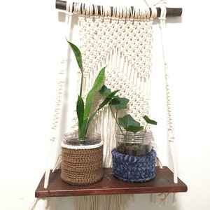 May include: A macrame wall hanging with a wooden dowel and a shelf underneath. Two plants are in glass jars with crocheted covers, one brown and one blue.