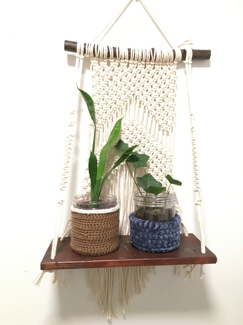 May include: A macrame wall hanging with a wooden dowel and a shelf underneath. Two plants are in glass jars with crocheted covers, one brown and one blue.