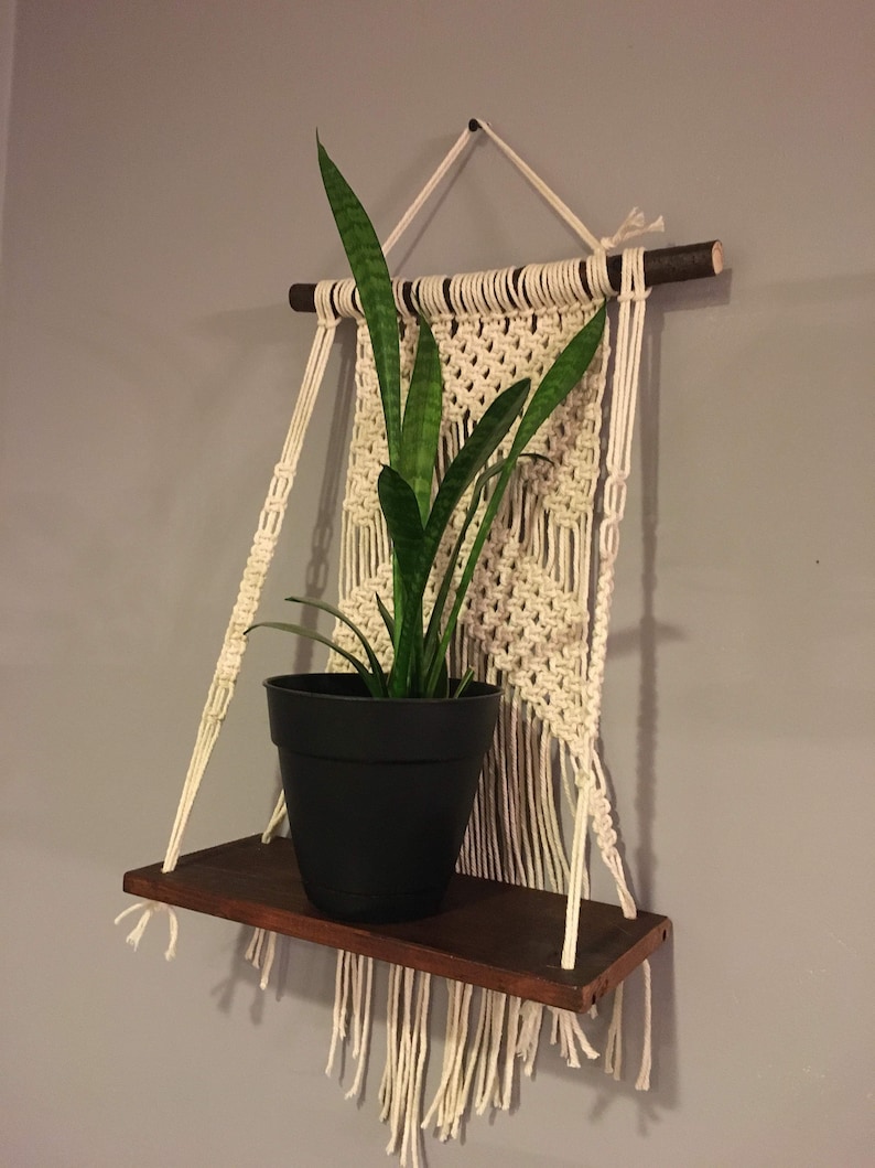 May include: A macrame wall hanging with a wooden shelf and a potted plant. The macrame is made with white cord and has a fringe at the bottom. The shelf is made of dark wood and is attached to the macrame with two cords. The plant is a snake plant in a black pot.