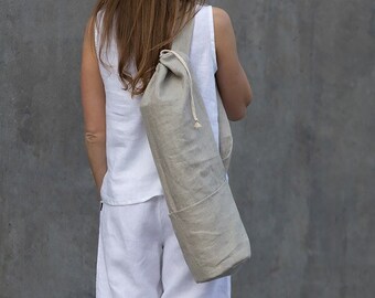 May include: A person wearing white pants and a white sleeveless top carries a light beige linen yoga mat bag over their shoulder.