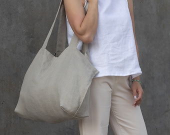 May include: A large, light gray linen tote bag with two wide shoulder straps. The bag features a simple, unstructured design with a spacious main compartment and a rectangular base, crafted from a textured, natural-looking fabric.