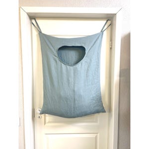 May include: A pale blue fabric laundry bag with a large oval opening, suspended from a white door. The bag hangs by thin straps and has a gathered base. The door features a silver doorknob.