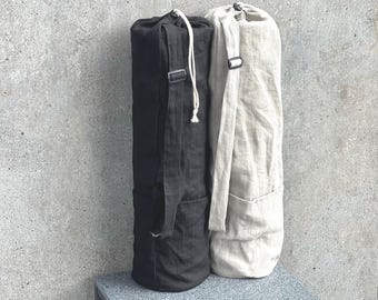 May include: Two cylindrical yoga mat bags. One is black, the other is a light beige. Both have a drawstring closure at the top and a shoulder strap. The black bag has a white rope drawstring, and the beige bag has a matching beige strap.