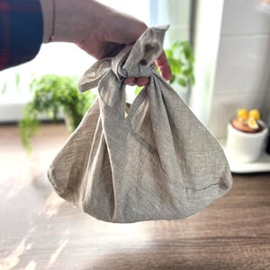 Puede incluir: Una bolsa de compras reutilizable de lino beige con un asa anudada. La bolsa está doblada y se sostiene en una mano.