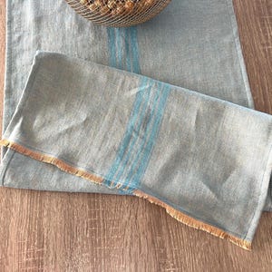 May include: A pale blue linen table runner with a fringed edge. The runner has vertical stripes of a lighter blue. The fabric is folded over on the bottom, and the runner is placed on a wooden surface.