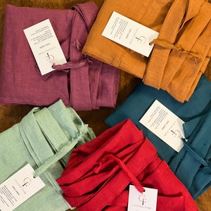 May include: An assortment of folded linen robes in various colors, including red, teal, orange, purple, and green. Each robe has a matching tie and a tag that reads "GOLDEN FLAX 100% LINEN". The robes are neatly arranged on a wooden surface.