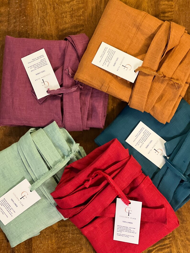 May include: An assortment of folded linen robes in various colors, including red, teal, orange, purple, and green. Each robe has a matching tie and a tag that reads "GOLDEN FLAX 100% LINEN". The robes are neatly arranged on a wooden surface.