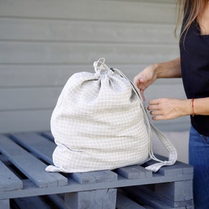 May include: A beige drawstring bag with a white checkered pattern, sitting on a grey wooden pallet. The bag has long straps and is made of a soft, woven fabric.