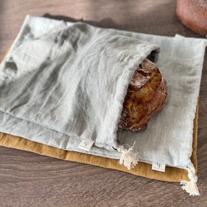 Può includere: Una pagnotta di pane parzialmente all'interno di un sacchetto di pane di lino grigio chiaro, appoggiata su un sacchetto di lino color senape. Le borse hanno etichette bianche con testo e un nappa. Un'altra pagnotta di pane è visibile sullo sfondo.