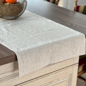 Può includere: Un runner da tavolo in lino beige con sottili strisce marroni. Il runner è appoggiato su un tavolo di legno.