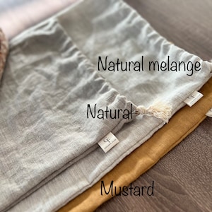 Op de afbeelding: Drie linnen broodzakken in verschillende kleuren: naturel, natuurlijke melange en mosterd. De zakken zijn gevouwen en op elkaar gestapeld. De zakken hebben een trekkoord sluiting en een klein label met de letter "G" erop.