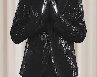 May include: A black blazer covered in reflective black sequins with a solid black lapel, worn over a black shirt and paired with black trousers. The garment features a button closure and two side pockets, showcasing a dark, shimmering texture.