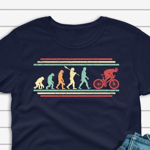 May include: Navy blue t-shirt featuring a retro-style evolution of man to cyclist graphic. The design includes figures in various stages of evolution, culminating in a cyclist on a red bicycle. The shirt is paired with blue jeans and white sneakers.