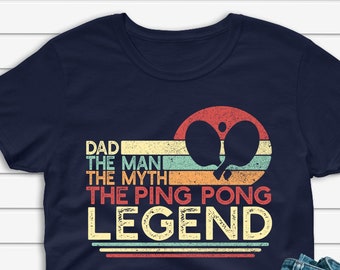 Könnte beinhalten: Marineblaues T-Shirt mit dem Text "DAD THE MAN THE MYTH THE PING PONG LEGEND" und einer Tischtennis-Grafik. Das Shirt wird mit blauen Jeans und weißen Turnschuhen kombiniert.