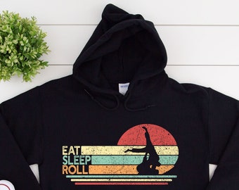 May include: Black hoodie with the text "EAT SLEEP ROLL" in a retro design. The graphic features a silhouette of a person doing a handstand against a sunset background. The hoodie is paired with white sneakers and blue jeans.